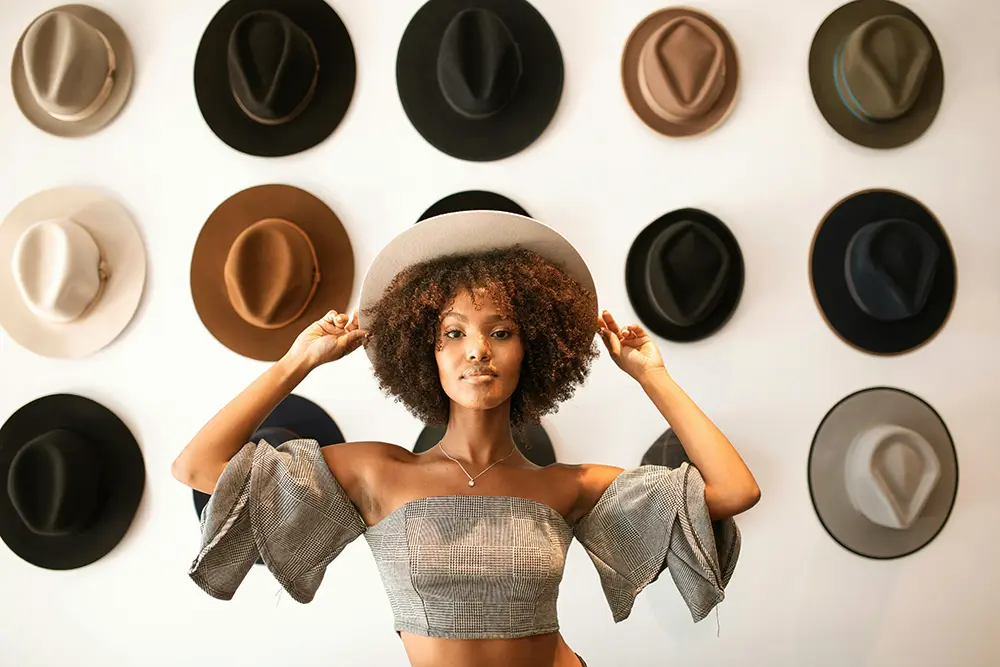 Photo d'une femme tenant son chapeau à deux mains devant un mur affichant plusieurs chapeaux.